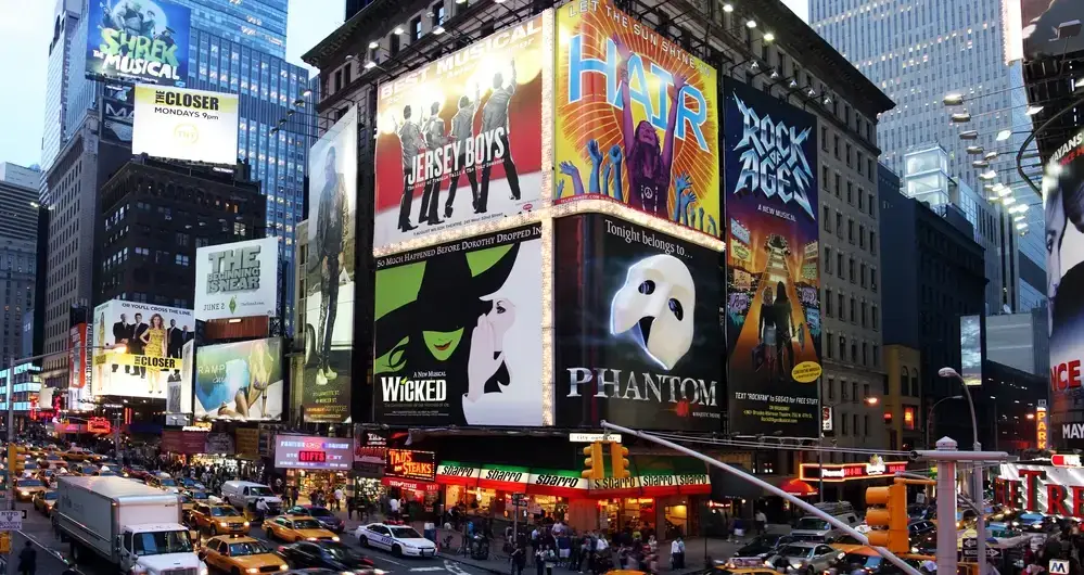 Brightly lit Broadway billboards for musicals like Wicked and Phantom, with heavy traffic below exemplify attention biases in a busy city.