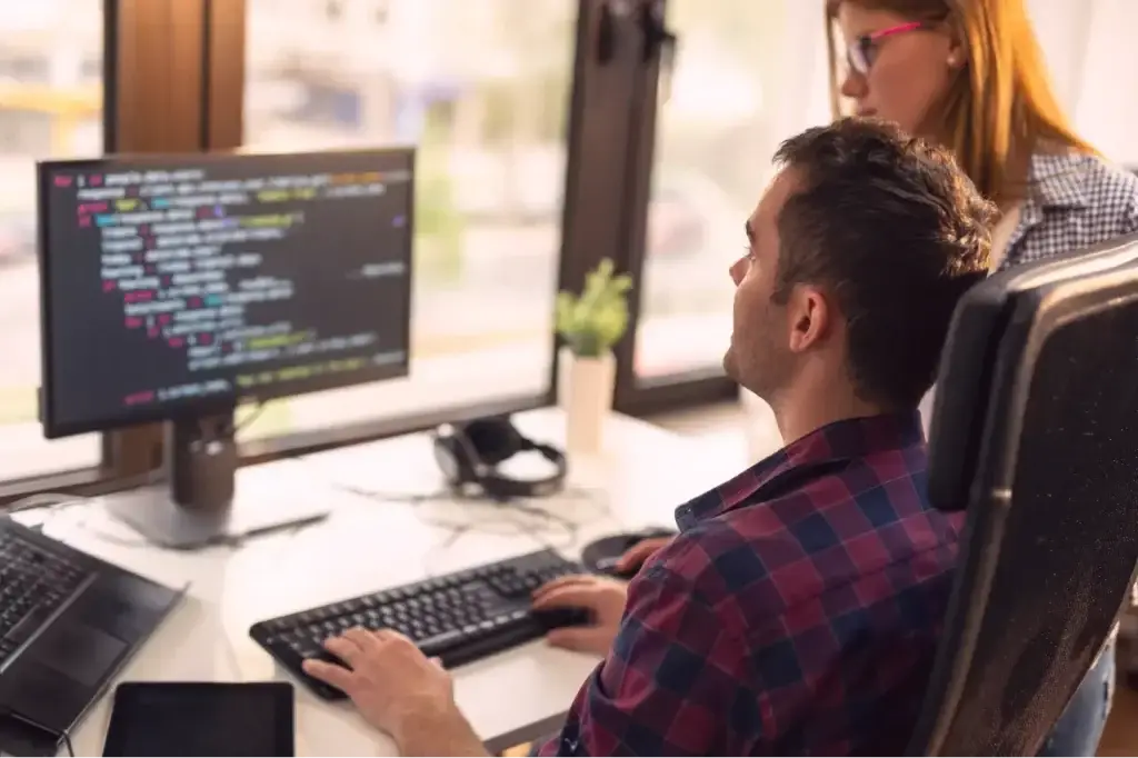 Two web developers coding a landing page; a man types on a keyboard while a woman stands behind him. Discusses how much does a landing page cost.