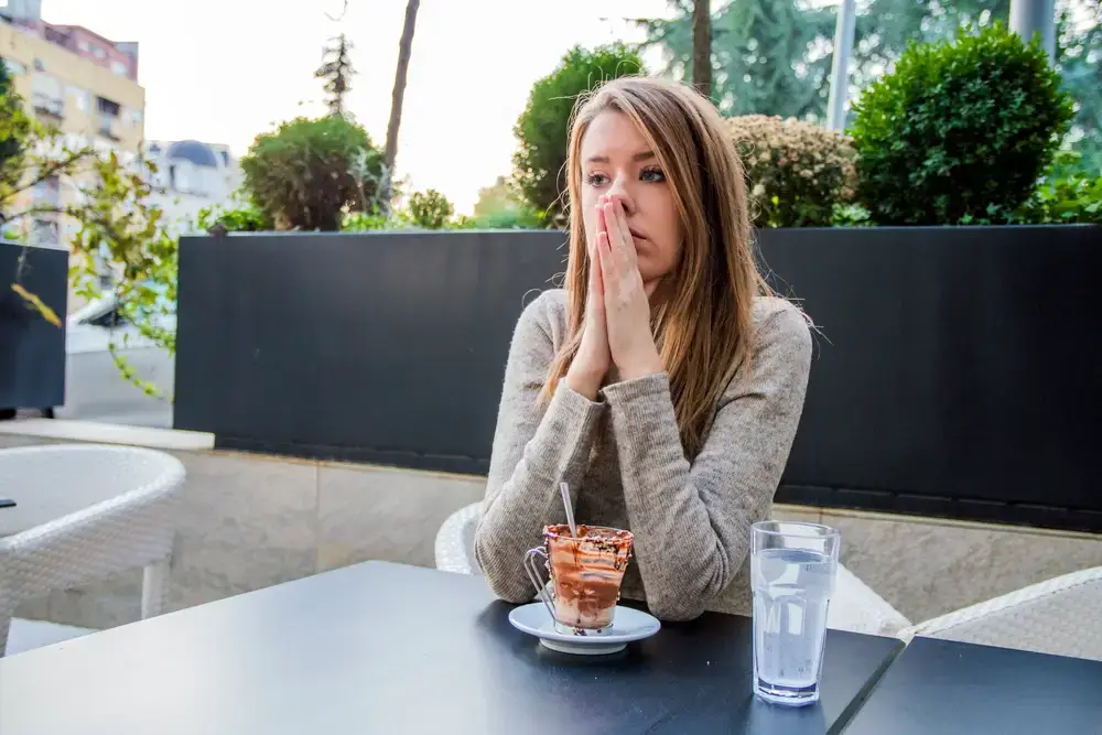 a woman sitting outside at a café, appearing stressed with her hands on her face, possibly contemplating digital marketing consultant strategies over a half-eaten dessert.